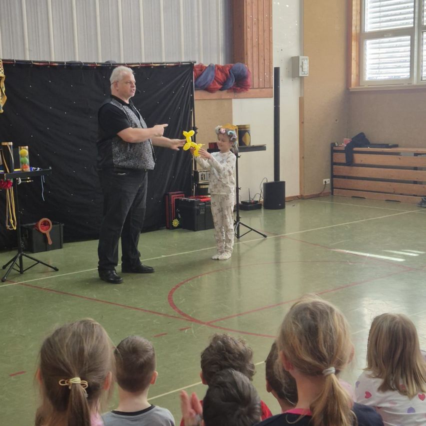 Ein Mann führt einen Ballontrick vor einem kleinen Mädchen und einem Publikum aus Kindern vor.
