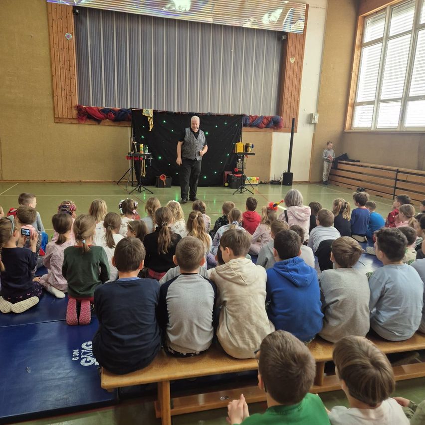 Ein Zauberer führt eine Show auf der Bühne in einer Turnhalle vor, während eine Gruppe von Kindern aufmerksam von einer Bank aus zusieht. Die Bühne ist mit bunten Dekorationen geschmückt.
