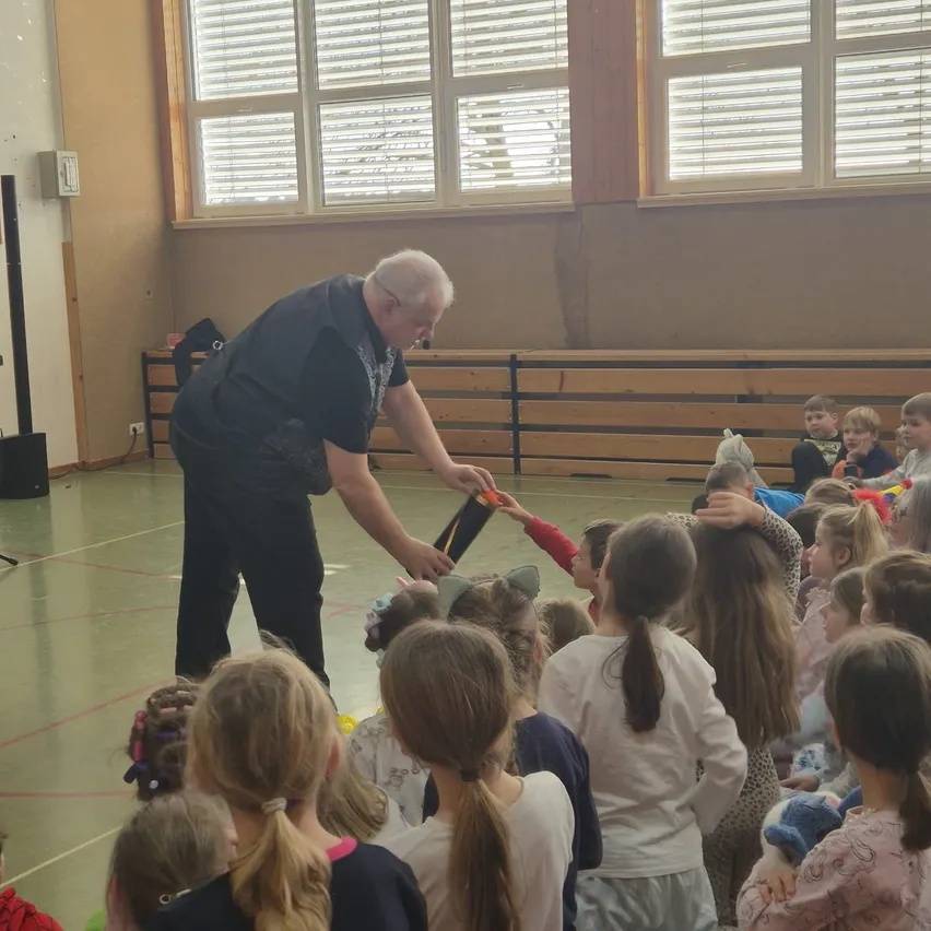 Ein Mann in einem schwarzen Shirt verteilt Spielzeug an eine Gruppe von Kindern in einer Turnhalle. Die Kinder sind um ihn herum versammelt und warten gespannt auf ihre Reihe. Die Turnhalle hat eine Holzbank, einen Lautsprecher und Fenster mit Jalousien.