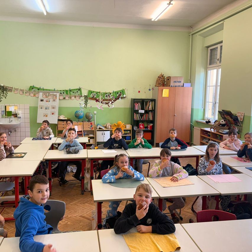Ein Klassenzimmer mit Kindern, die an ihren Schreibtischen Donuts essen, grüne Wände und Regale mit Büchern und einer Globus.