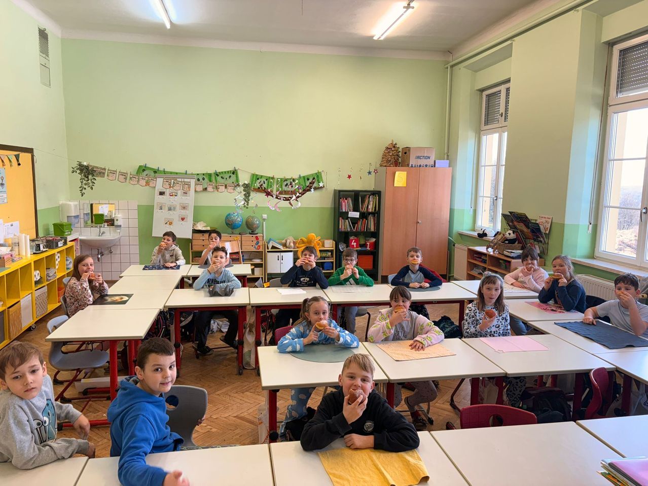 Ein Klassenzimmer mit Kindern, die an ihren Schreibtischen Donuts essen, grüne Wände und Regale mit Büchern und einer Globus.
