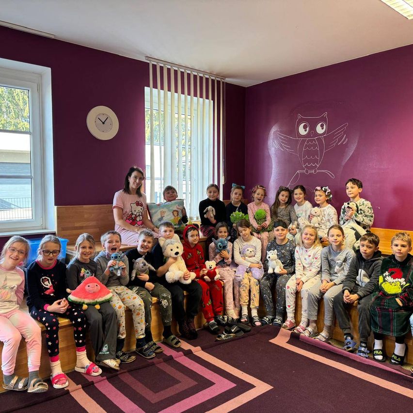 Eine Gruppe von Kindern und eine erwachsene Frau sitzen auf einer Bank in einem Raum mit lila Wänden. Einige tragen Pyjamas und halten Kuscheltiere. Eine Uhr und eine Eule sind an der Wand angebracht.