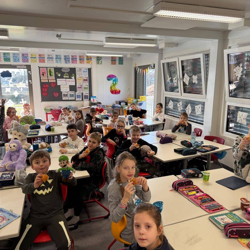 Ein Klassenzimmer voller Kinder, die Sandwiches und Snacks essen. Der Raum hat Schreibtische, Stühle und Poster an der Wand. Einige Kinder tragen Brillen.
