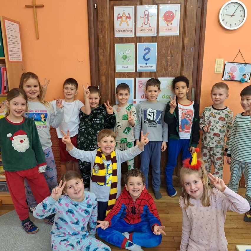 Gruppe von Kindern in Pyjamas posiert für ein Foto im Klassenzimmer, mit Lernplakaten und einer Uhr an der Wand.