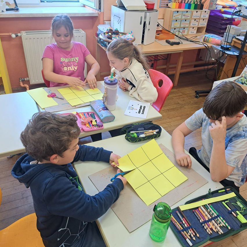 Eine Gruppe von Kindern sitzt an einem Tisch und arbeitet an einem Bastelprojekt. Ein Junge benutzt eine Schere, während andere Papiere und Werkzeuge um sich herum haben.