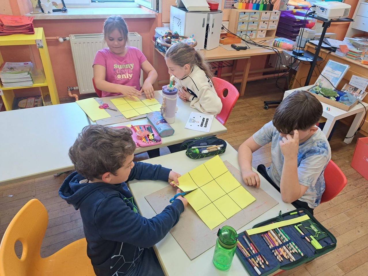 Eine Gruppe von Kindern sitzt an einem Tisch und arbeitet an einem Bastelprojekt. Ein Junge benutzt eine Schere, während andere Papiere und Werkzeuge um sich herum haben.