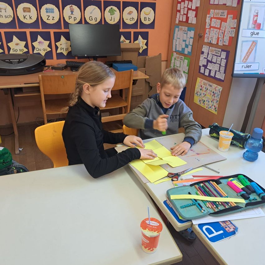 Zwei Kinder sitzen an einem Schreibtisch in einem Klassenzimmer und beschäftigen sich mit einer Bastelaktivität mit Schere, Papier und einer Flasche Kleber. Dahinter steht ein Schreibtisch mit einem Computer und einem Drucker.