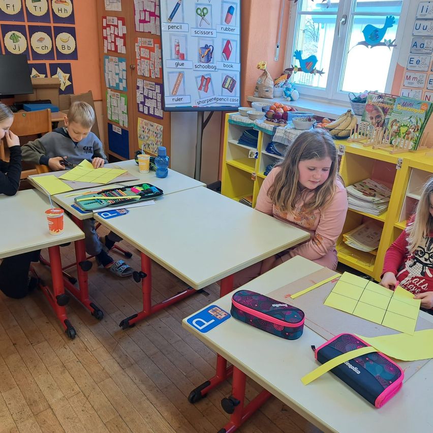 In einem Klassenzimmer sitzen mehrere Kinder an Tischen mit Papieren und Schulmaterial. Ein Kind hält ein Papier, ein anderes schaut in ein Buch. An der Wand hängen Poster, ein Regal mit Bananen und ein Fenster mit Blick nach draußen.