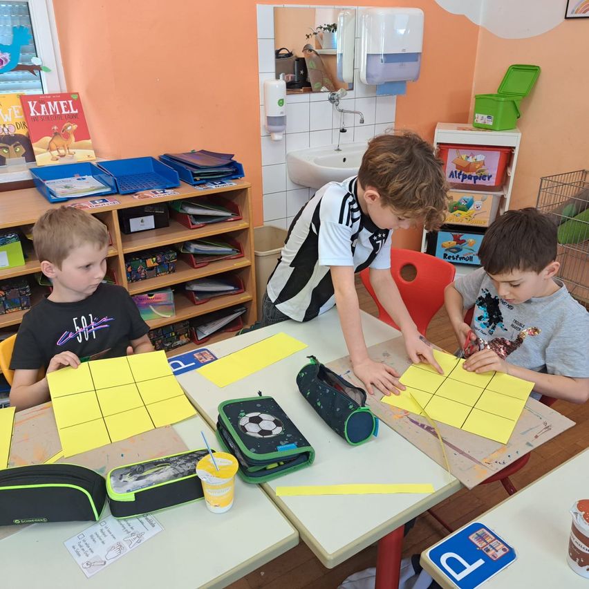 Drei Jungen sitzen in einem Klassenzimmer an einem Tisch und sind mit einer Papierschneideaktivität beschäftigt. Ein Junge benutzt eine Schere, während die anderen beiden zusehen. Der Tisch ist mit Papier, Scheren und anderen Materialien übersät. Hinter ihnen ist ein Schrank mit Büchern und Aufbewahrungsboxen zu sehen, und ein Spiegel reflektiert einen Handdesinfektionsspender.