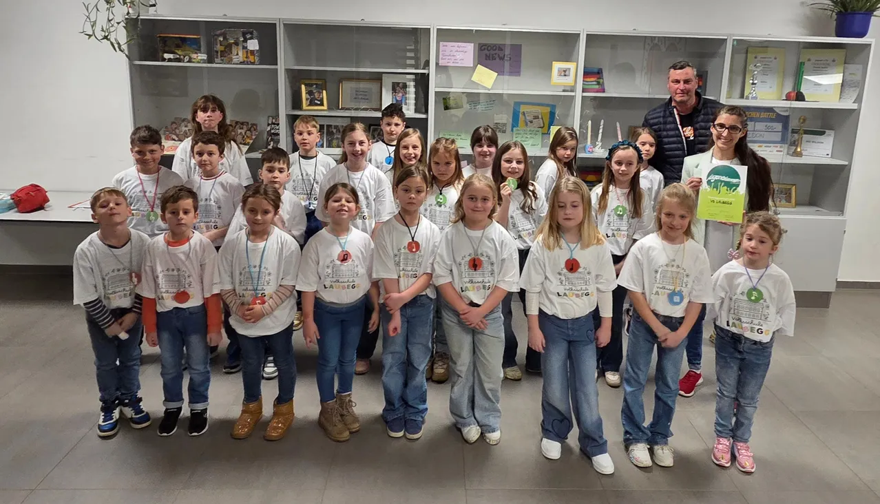 Eine Gruppe von Kindern, wahrscheinlich Schüler, in weißen T-Shirts mit Abzeichen, steht in einem Klassenzimmer mit Regalen im Hintergrund.