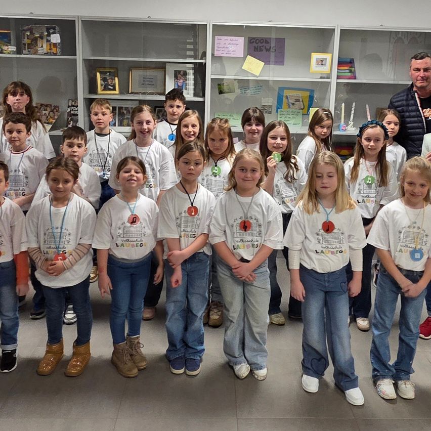Eine Gruppe von Kindern, wahrscheinlich Schüler, in weißen T-Shirts mit Abzeichen, steht in einem Klassenzimmer mit Regalen im Hintergrund.