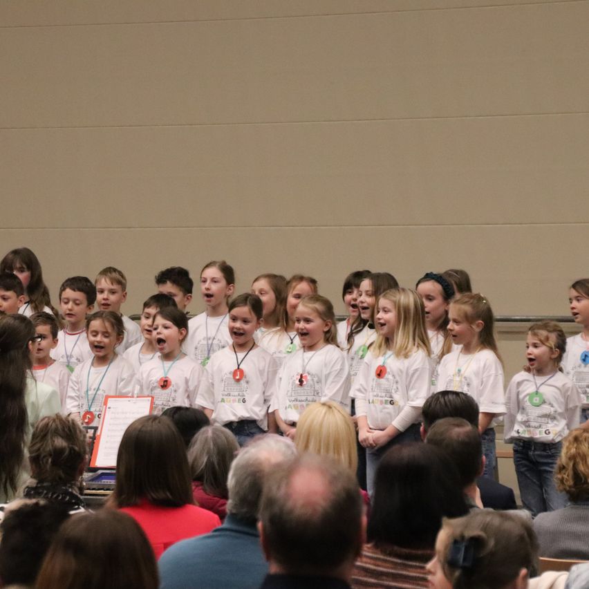 Ein Chor von Kindern in weißen Hemden mit Abzeichen tritt vor einem Publikum in einem Saal auf.