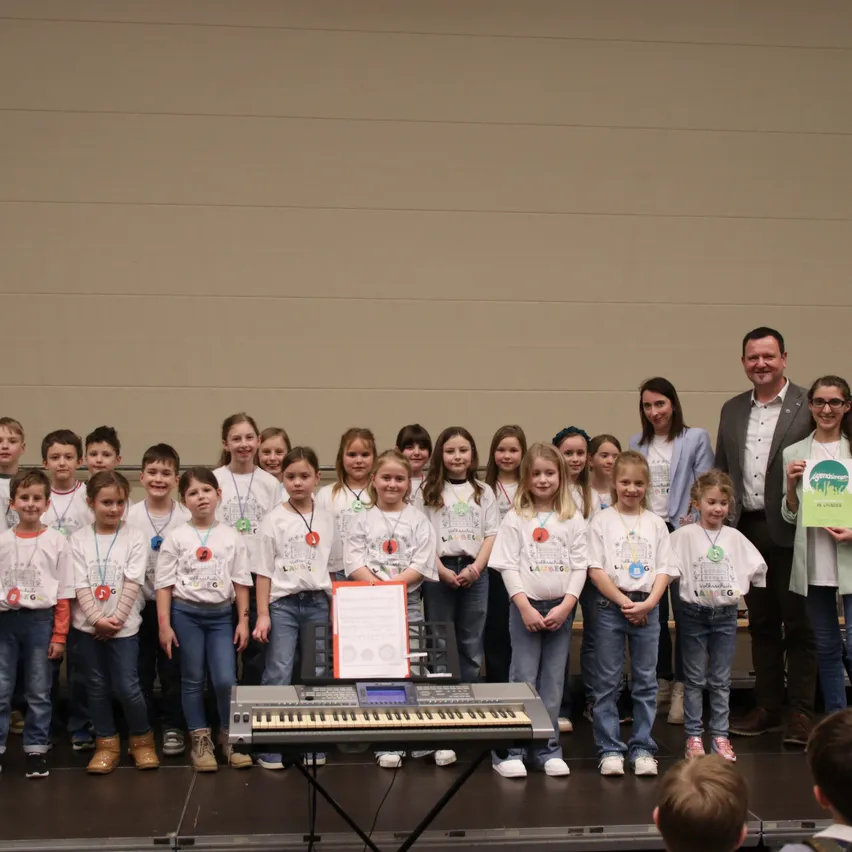 Eine Gruppe von Kindern mit weißen T-Shirts und Namensschildern steht mit Erwachsenen auf der Bühne, hält ein Zertifikat und posiert für ein Foto. Ein Keyboard steht vor ihnen.