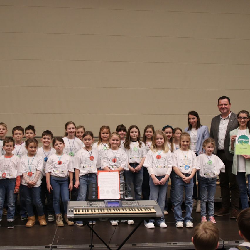 Eine Gruppe von Kindern mit weißen T-Shirts und Namensschildern steht mit Erwachsenen auf der Bühne, hält ein Zertifikat und posiert für ein Foto. Ein Keyboard steht vor ihnen.