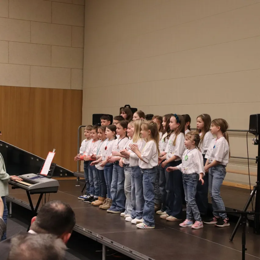 Ein Chor junger Kinder tritt mit einem Pianisten auf der Bühne vor einem Publikum auf.
