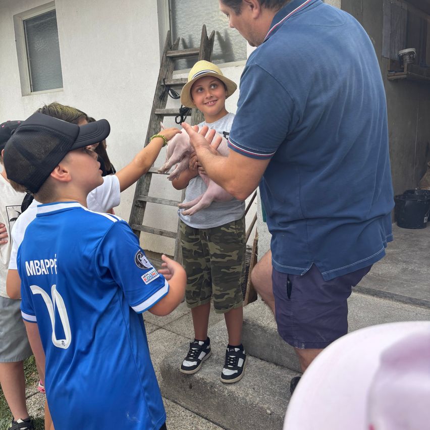 Ein Mann hält ein Ferkel, während ein Junge in einem blauen Mbappe-Trikot und ein anderes Kind es berühren wollen. Eine Leiter und ein Gebäude sind im Hintergrund.