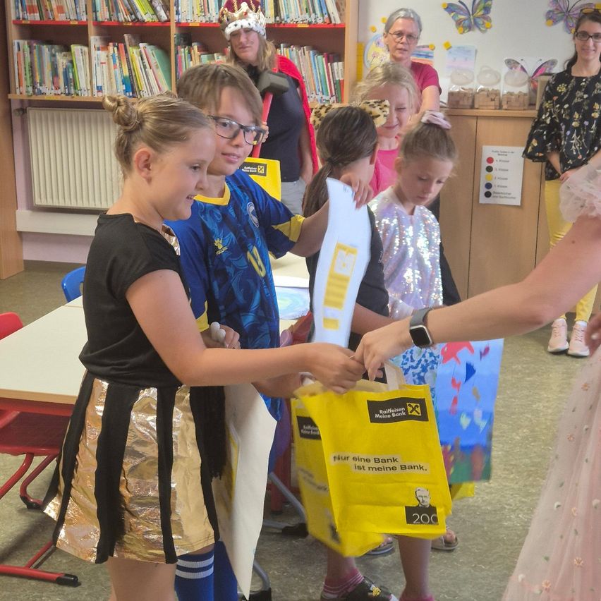 Eine Gruppe von Kindern in einem Klassenzimmer, eines hält einen gelben Beutel, ein anderes eine weiße Papier, während eine Frau Taschen verteilt.