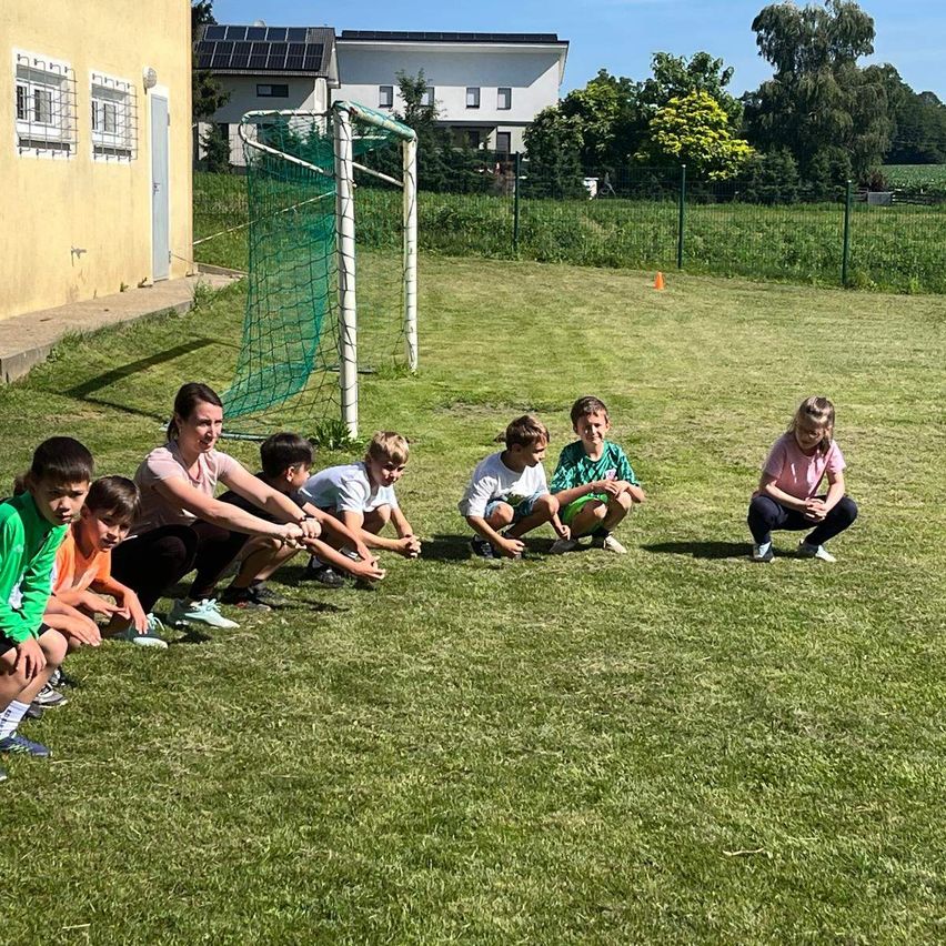 Eine Gruppe von Kindern und einer erwachsenen Frau sitzen auf einem Rasen, möglicherweise bei einer Übung. Hinter ihnen steht ein Fußballtor, und in der Ferne befinden sich Häuser und Bäume.