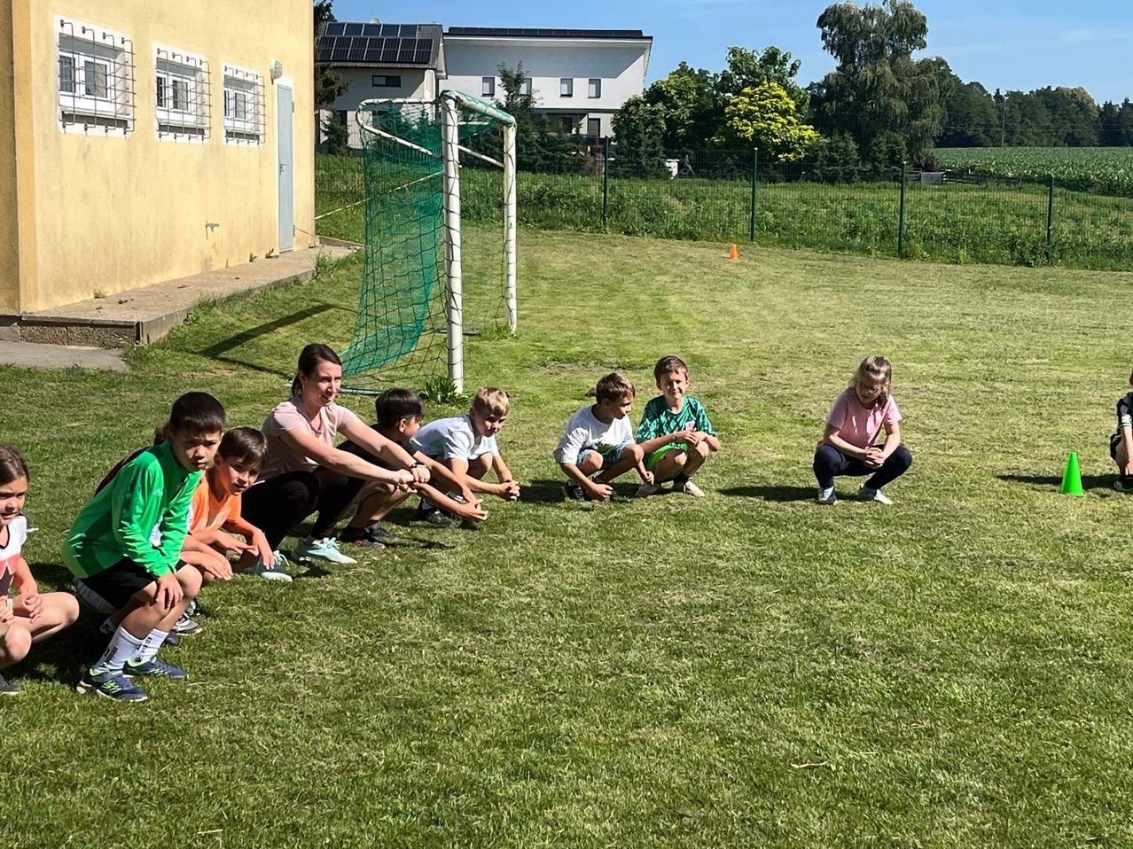 Eine Gruppe von Kindern und einer erwachsenen Frau sitzen auf einem Rasen, möglicherweise bei einer Übung. Hinter ihnen steht ein Fußballtor, und in der Ferne befinden sich Häuser und Bäume.