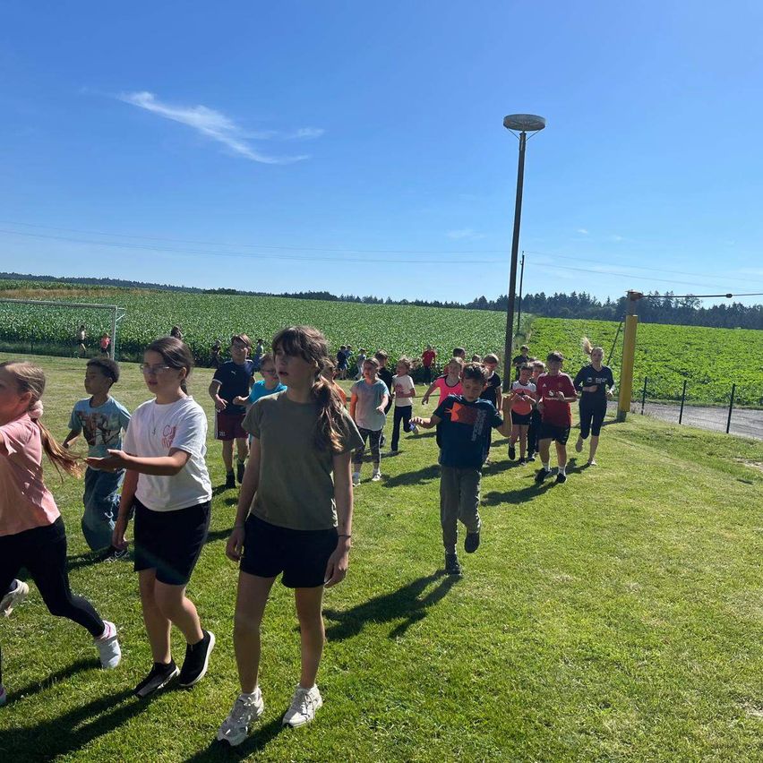 Eine Gruppe von Kindern läuft auf einem Rasenfeld, wahrscheinlich bei einer Schulsportveranstaltung. Sie tragen Turnschuhe und einige tragen eine Brille. In der Ferne laufen Menschen auf der Straße. Es gibt auch Bäume und ein Feld mit Pflanzen.
