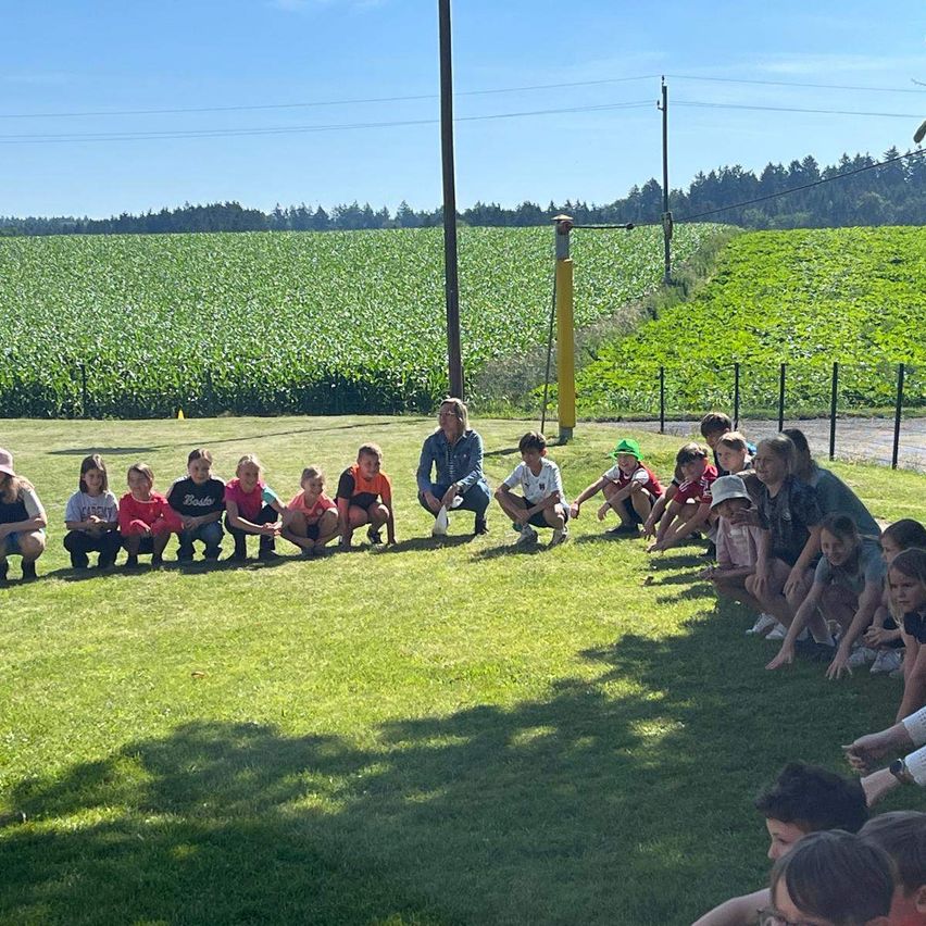Eine Gruppe von Kindern und Erwachsenen sitzt in einer Reihe auf einem Grasfeld, mit einer Frau vor ihnen. Dahinter befindet sich ein Feld mit Pflanzen und ein Zaun.