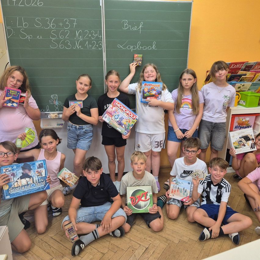 Eine Gruppe von Kindern posiert für ein Foto, während sie verschiedene Brettspiele und Spielzeug in den Händen halten. Sie befinden sich in einem Klassenzimmer mit einer Tafel und einem Bücherregal im Hintergrund.