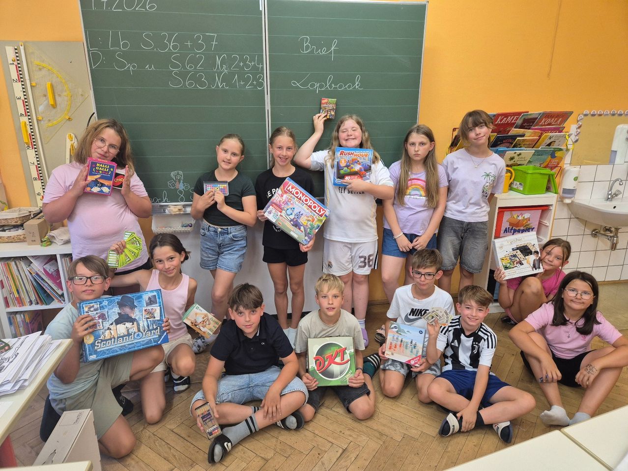 Eine Gruppe von Kindern posiert für ein Foto, während sie verschiedene Brettspiele und Spielzeug in den Händen halten. Sie befinden sich in einem Klassenzimmer mit einer Tafel und einem Bücherregal im Hintergrund.