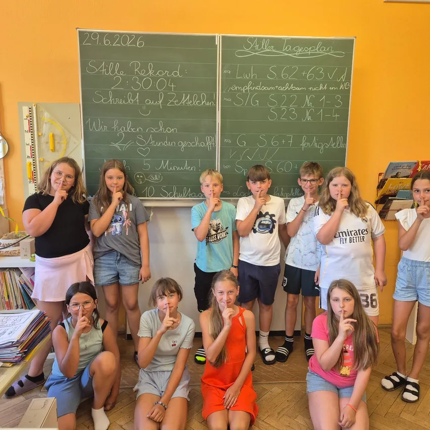 Eine Gruppe von Kindern in einem Klassenzimmer posiert für ein Foto mit den Fingern auf den Lippen. Dahinter zeigen zwei Tafeln verschiedene mathematische Gleichungen und Aufzeichnungen.