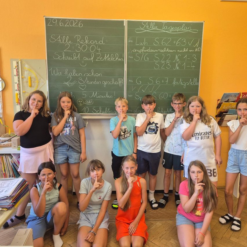 Eine Gruppe von Kindern in einem Klassenzimmer posiert für ein Foto mit den Fingern auf den Lippen. Dahinter zeigen zwei Tafeln verschiedene mathematische Gleichungen und Aufzeichnungen.