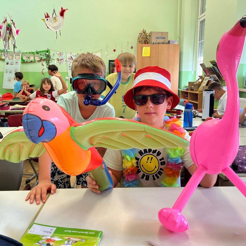 Zwei Jungen in einem Klassenzimmer tragen aufblasbare Tierkostüme, einer hält einen Papagei und der andere einen Flamingo. Sie lächeln und posieren für ein Foto.