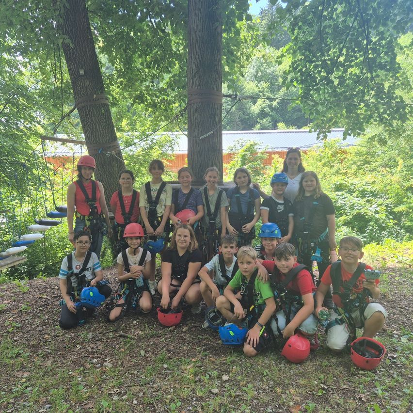 Eine Gruppe von Kindern und Erwachsenen in Sicherheitsausrüstung posiert für ein Foto in einer Waldumgebung mit Bäumen und einer Seilrutsche im Hintergrund.