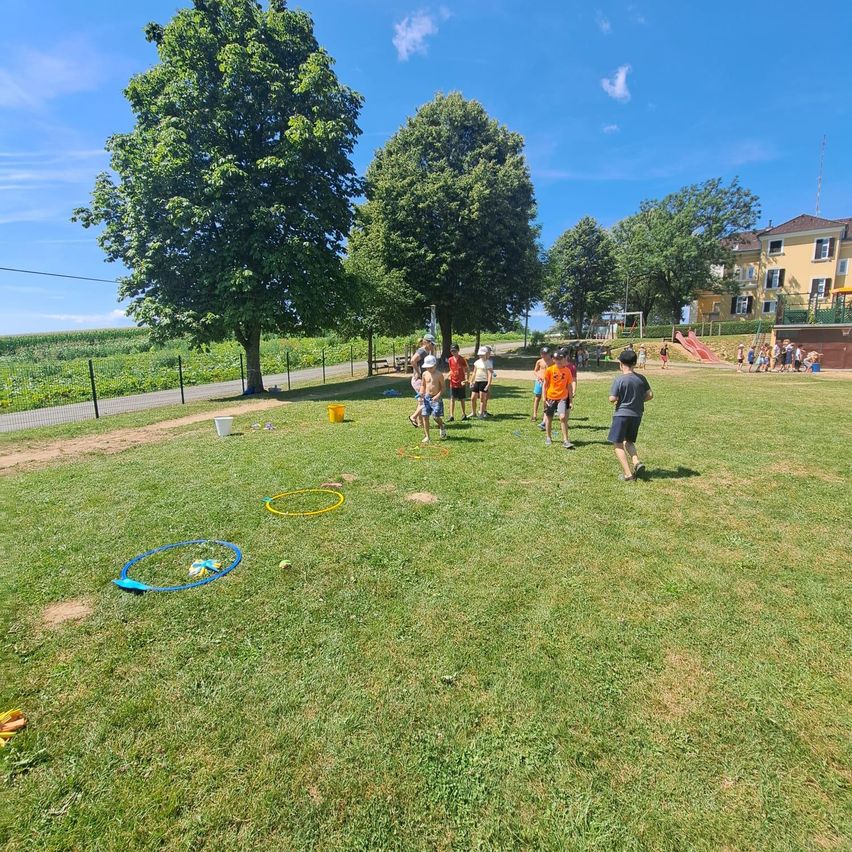 Eine Gruppe von Kindern und Erwachsenen spielt auf einem Rasenfeld mit Hula-Hoops und Eimern. Bäume und ein Gebäude sind im Hintergrund.
