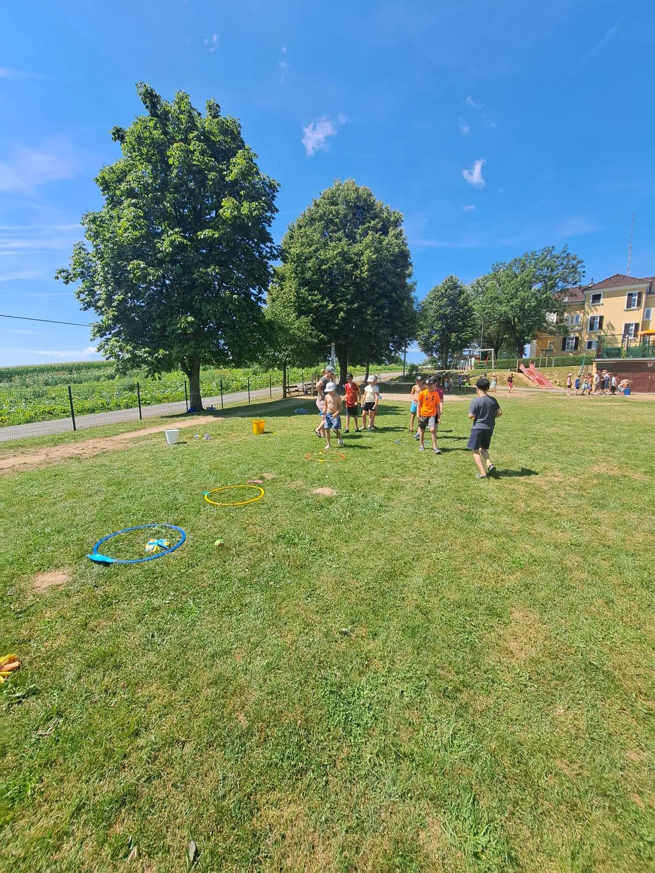 Eine Gruppe von Kindern und Erwachsenen spielt auf einem Rasenfeld mit Hula-Hoops und Eimern. Bäume und ein Gebäude sind im Hintergrund.
