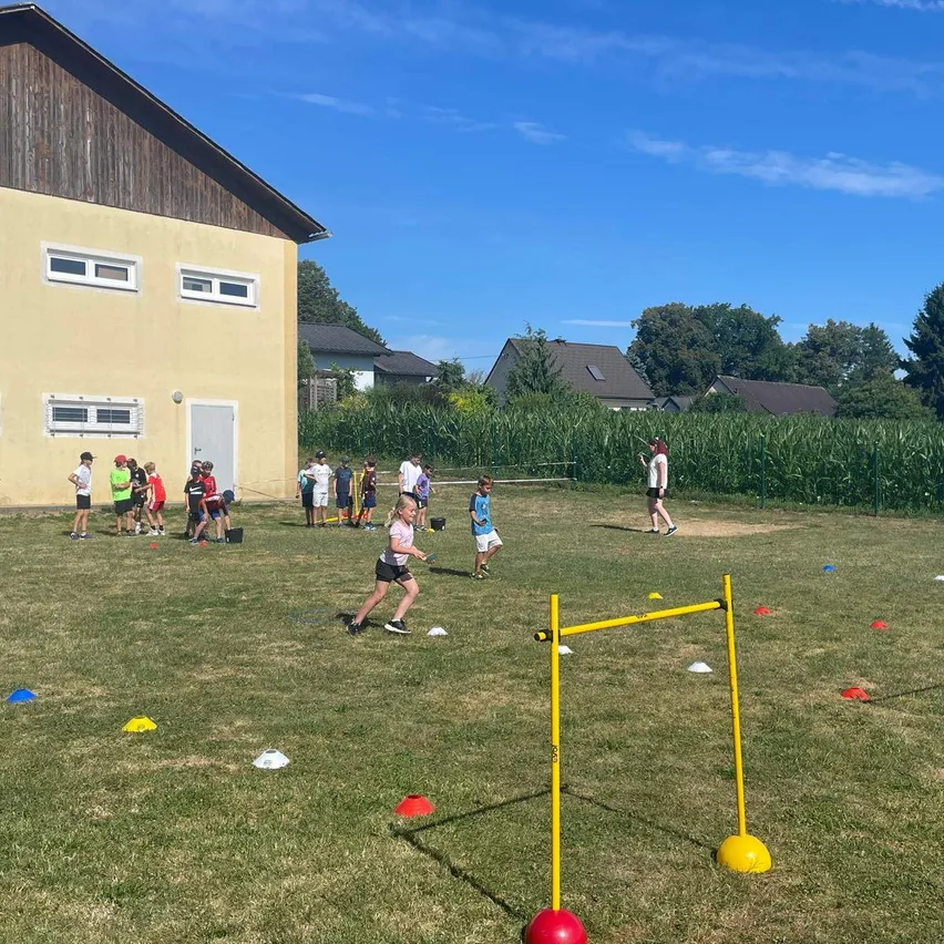 Eine Gruppe von Kindern spielt ein Spiel auf einem Grasfeld mit einem gelben Hindernisparcours. Einige laufen, während andere stehen und zuschauen.