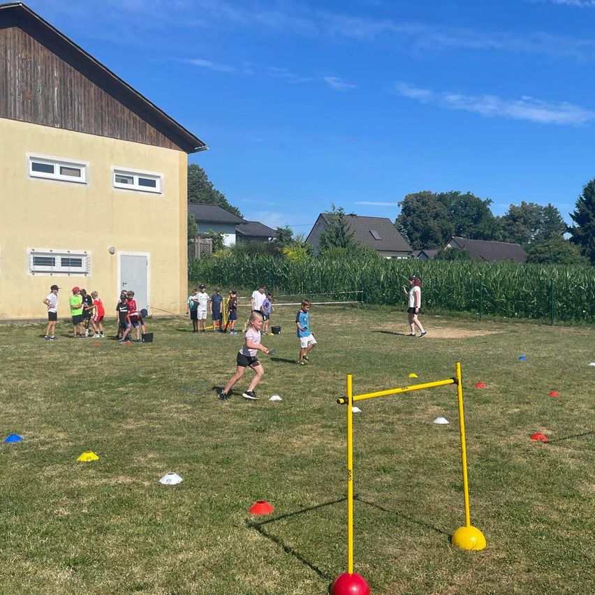 Eine Gruppe von Kindern spielt ein Spiel auf einem Grasfeld mit einem gelben Hindernisparcours. Einige laufen, während andere stehen und zuschauen.