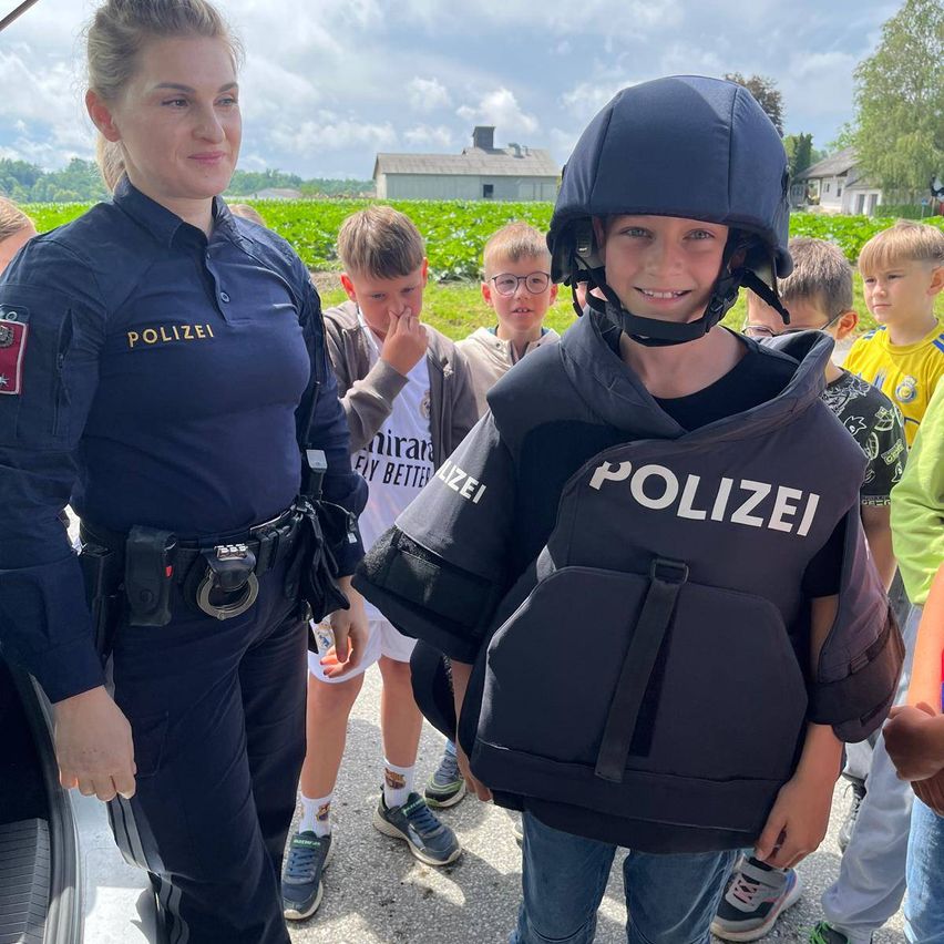 Eine Polizistin steht neben einem Jungen, der einen Polizeihelm und eine Weste trägt, mit mehreren Kindern im Hintergrund.