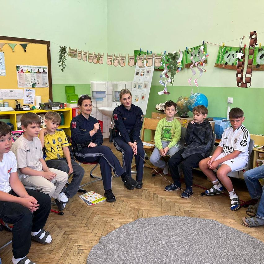Zwei Polizeibeamte sitzen mit einer Gruppe von Kindern in einem Klassenzimmer. Die Kinder hören aufmerksam zu. Es gibt einen Teppich und einen Globus auf dem Boden.