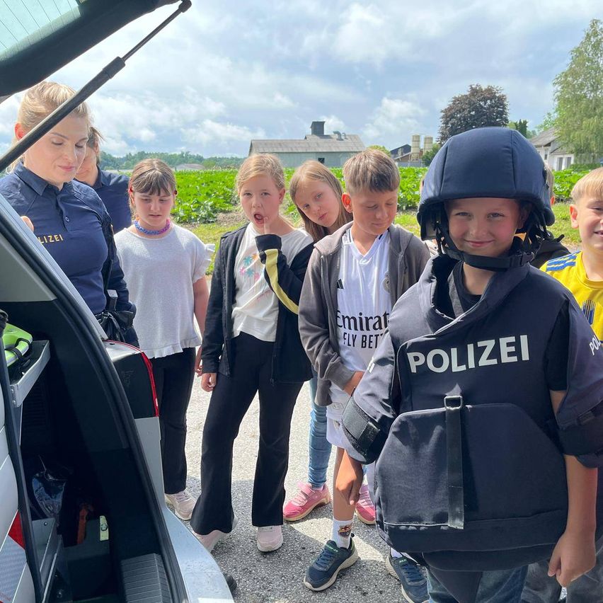 Kinder auf einem Feld mit einer Polizistin und einem Polizeiauto, ein Junge trägt eine Polizeiweste und einen Helm.