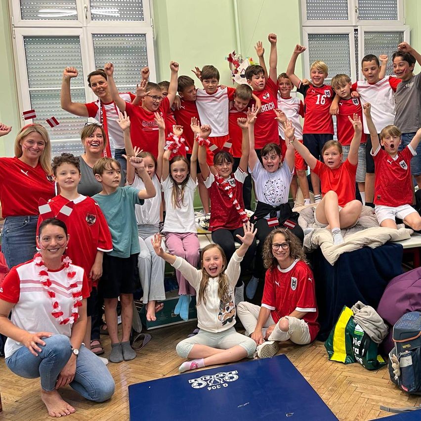 Eine Gruppe von Kindern und Erwachsenen in roten und weißen Trikots posiert für ein Foto in einem Raum mit Holzboden. Einige stehen und heben ihre Hände, während andere auf dem Boden sitzen.