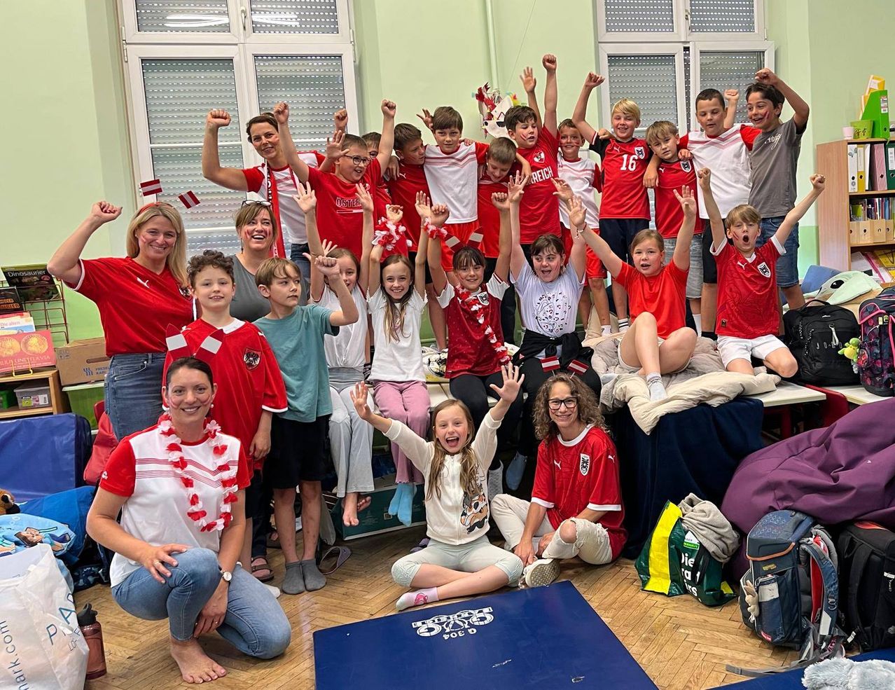 Eine Gruppe von Kindern und Erwachsenen in roten und weißen Trikots posiert für ein Foto in einem Raum mit Holzboden. Einige stehen und heben ihre Hände, während andere auf dem Boden sitzen.