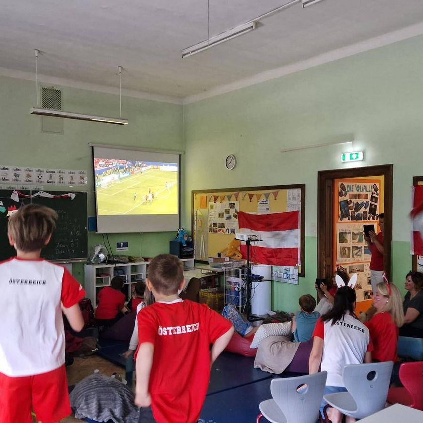 Ein Klassenzimmer mit einer Projektionsleinwand, die ein Fußballspiel zeigt. Schüler tragen rote und weiße Uniformen, und einige sitzen auf Beanbags. Eine Flagge und verschiedene Poster schmücken die Wände.