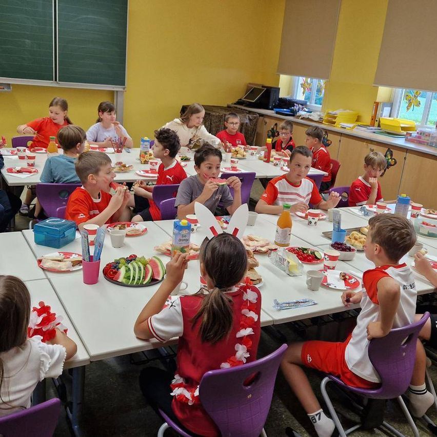 Mehrere Kinder in roten und weißen Uniformen sitzen an Tischen und essen eine Mahlzeit. Die Tische sind mit Tellern, Getränken und Snacks gedeckt. Die Kinder scheinen eine gute Zeit zu haben.