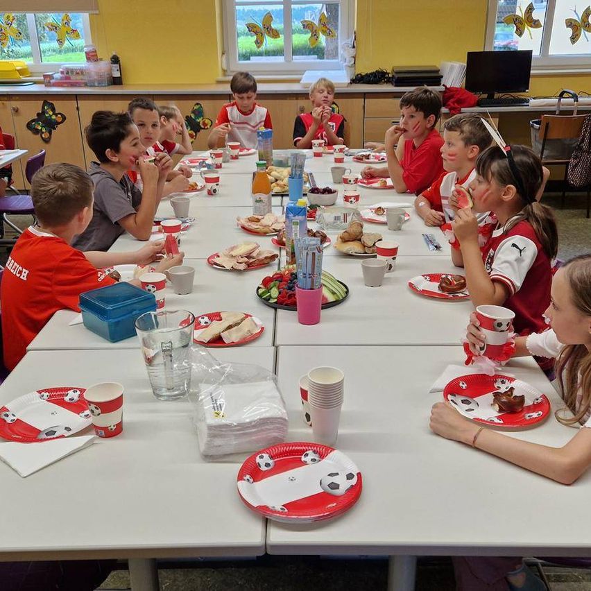 Kinder in Fußballtrikots sitzen um einen Tisch und essen. Der Tisch ist mit Tellern, Servietten und Getränken gedeckt. Der Raum hat gelbe Wände mit Schmetterlingsdekorationen.