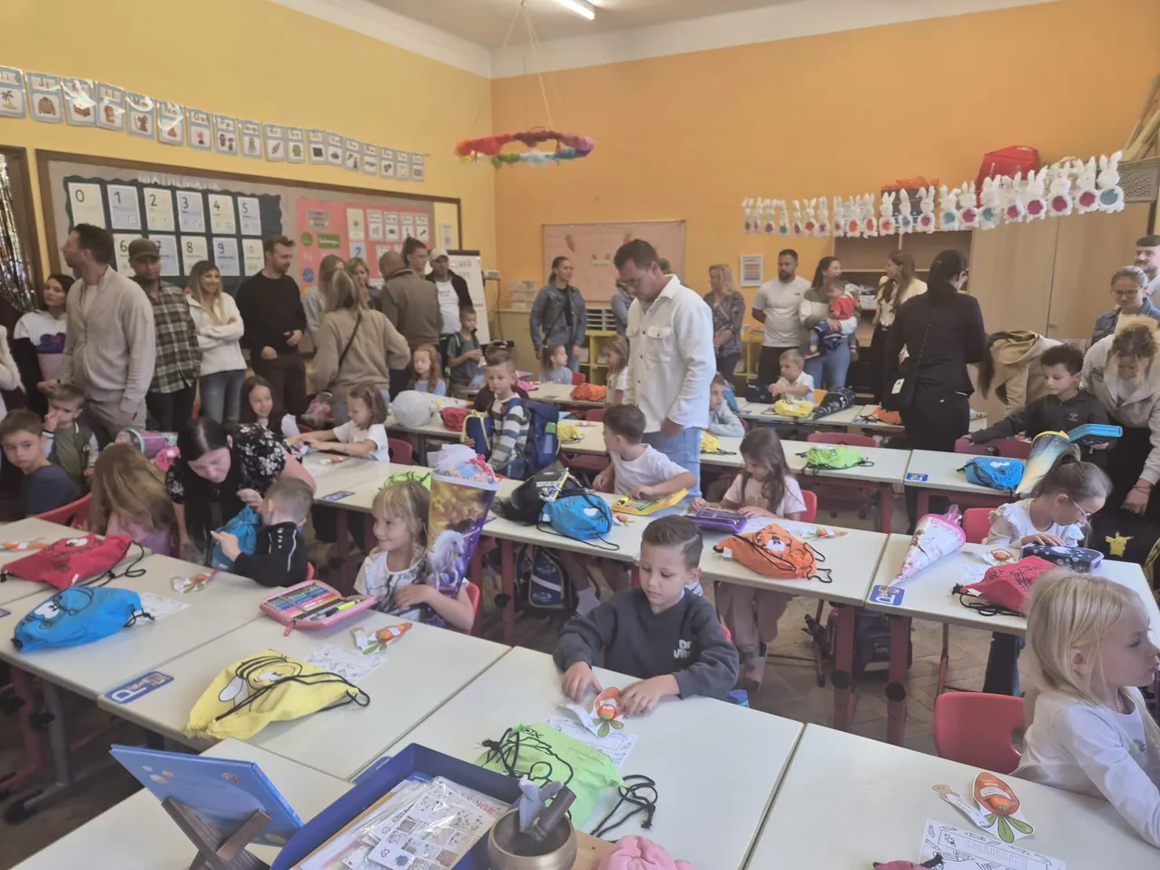 Ein Klassenzimmer voller Kinder und Erwachsener, einige stehen, einige sitzen und beschäftigen sich mit verschiedenen Aktivitäten.