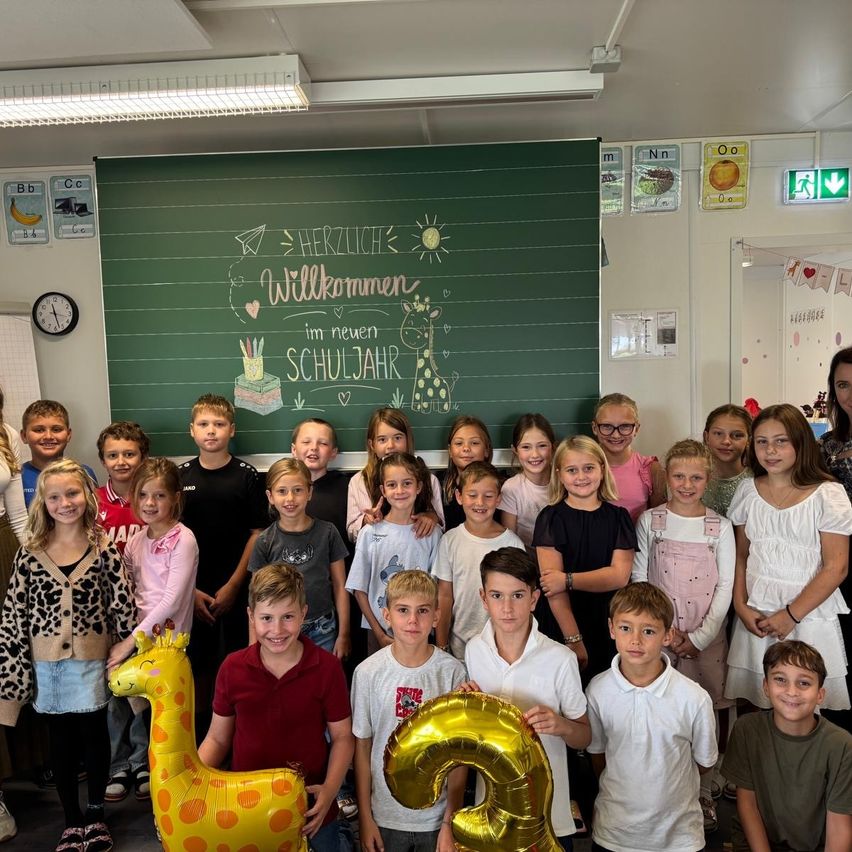 Eine Gruppe von Kindern und zwei Frauen posiert für ein Foto im Klassenzimmer. Die Tafel hinter ihnen hat eine Nachricht, die 'Herzlich Willkommen im neuen Schuljahr' lautet.