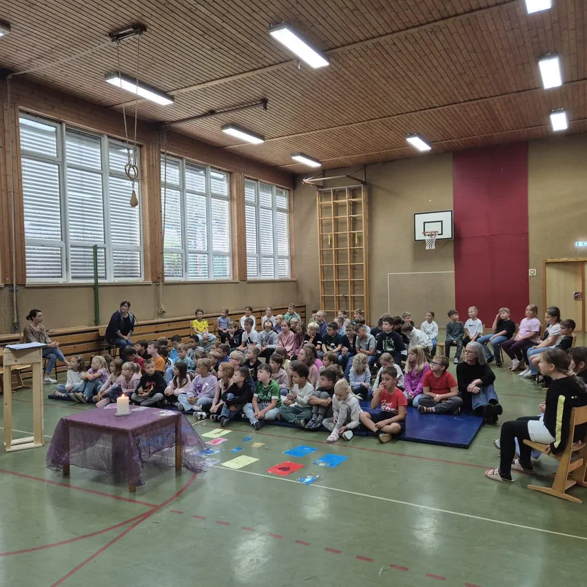 Ein Priester steht vor einer Gruppe von Kindern, die auf dem Boden in einer Turnhalle sitzen.