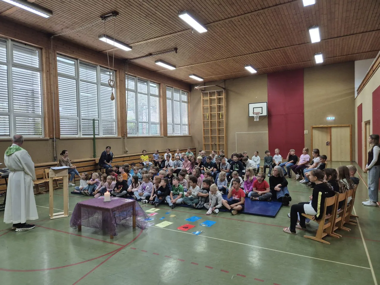 Ein Priester steht vor einer Gruppe von Kindern, die auf dem Boden in einer Turnhalle sitzen.