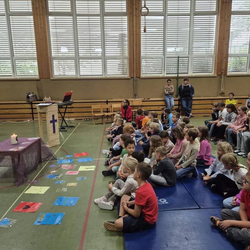 Eine Gruppe von Kindern sitzt auf dem Boden in einem Raum und hört einer Person zu, die vor einem Podium mit einem Kreuz darauf steht.