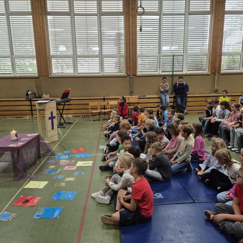 Eine Gruppe von Kindern sitzt auf dem Boden in einem Raum und hört einer Person zu, die vor einem Podium mit einem Kreuz darauf steht.