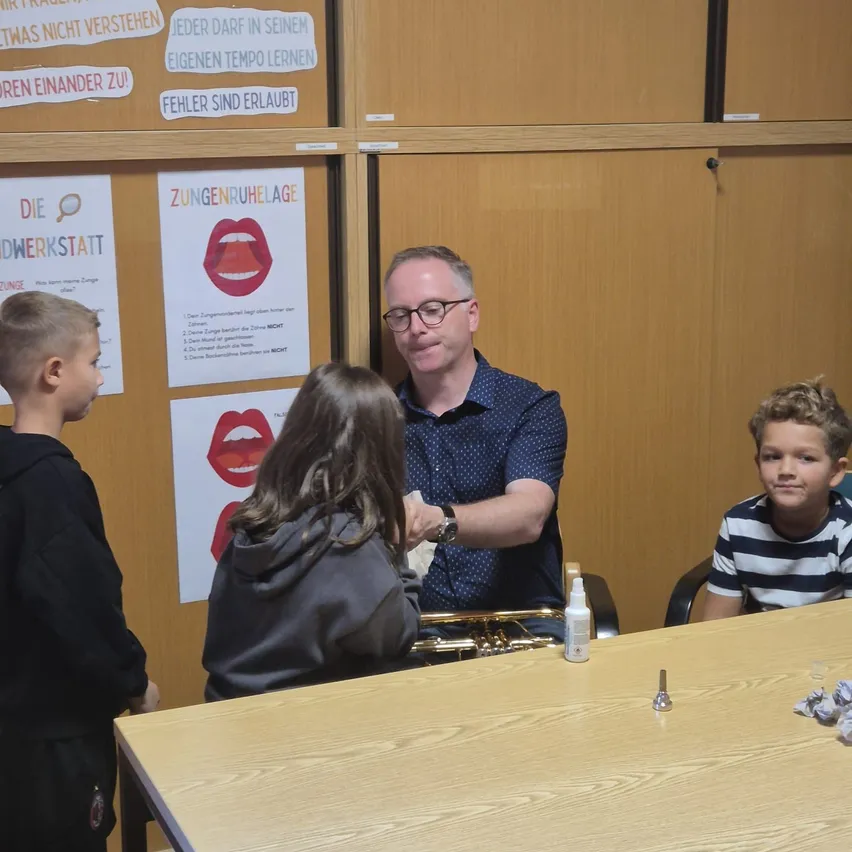 Ein Mann sitzt an einem Tisch mit drei Kindern. Er lächelt und hält ein Posaune. Die Kinder schauen ihn an und scheinen zuzuhören. Hinter ihnen ist eine Wand mit Plakaten.